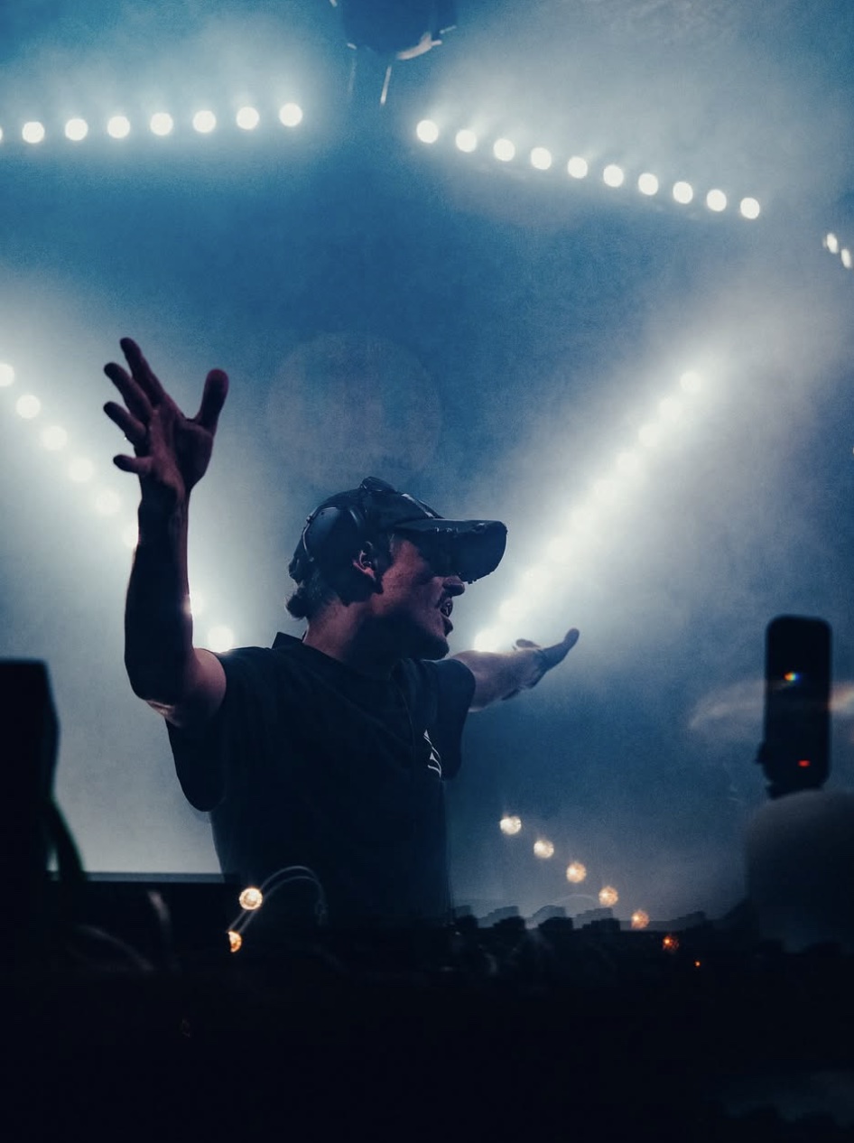 Dennis Reif behind the decks, arms outstretched beneath blue stage lights.